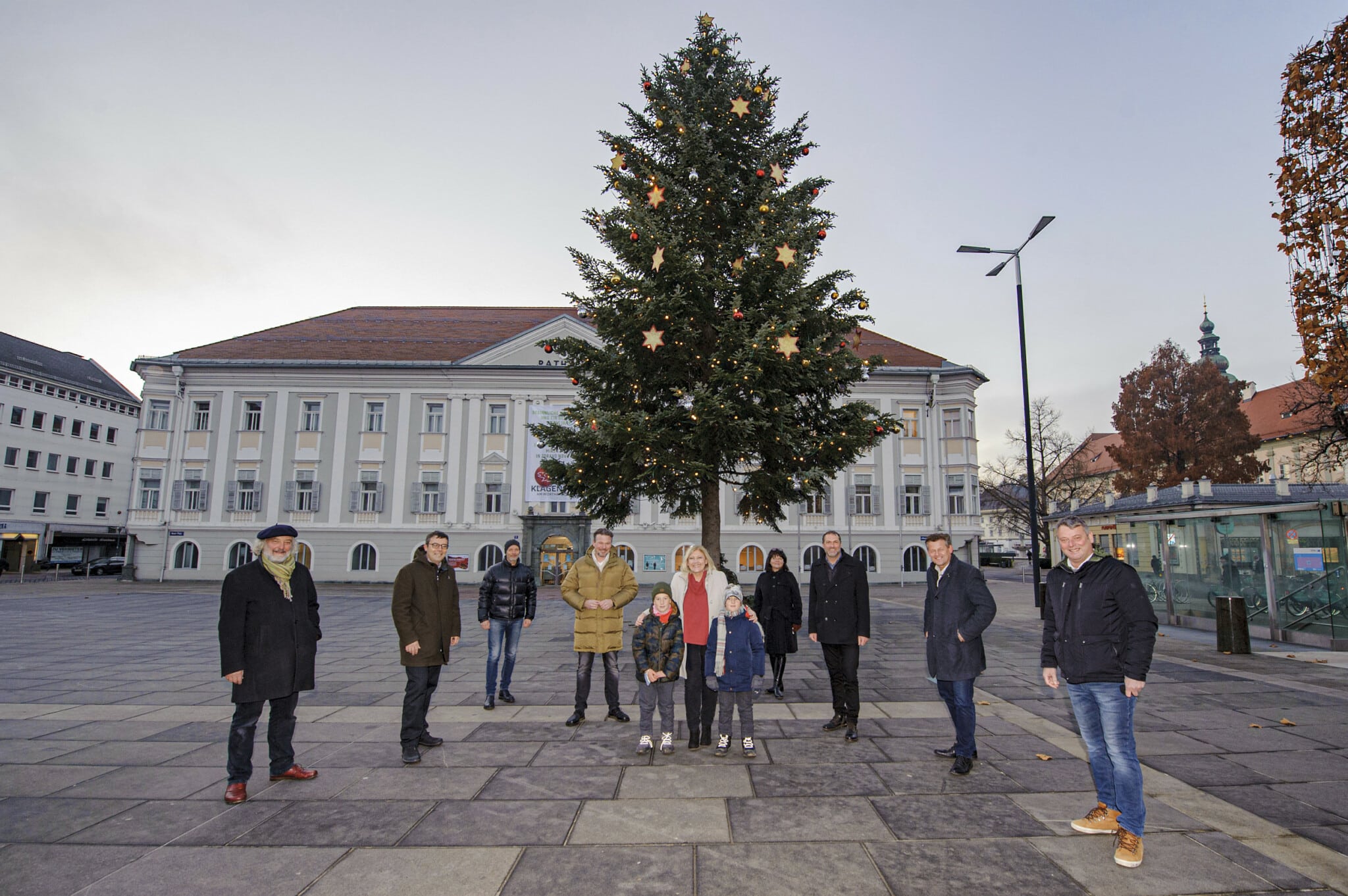 Vertreter des Stadtsenates und des Gemeinderates vor dem beleuchteten Christbaum am Neuen Platz. Neben der Bürgermeisterin stehen die beiden Kinder Leopold und Taddäus.